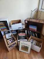 Full lot showing small wooden bookshelf, cardboard boxes with books and DVDs, vintage wrought iron page holder on top of bookshelf