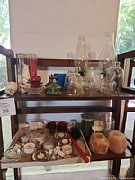 Wide view of wooden shelves holding various glass candle holders including hurricane lamps, red glass holders, oil lamp base, and multiple candles including salt candles and votives