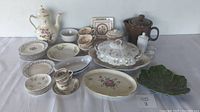 Full view of assorted china and pottery including plates, bowls, cups, serving dishes, teapots and decorative pieces arranged on a cloth.