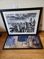 Two framed art pieces propped against wall on hardwood floor under lot tag 43. A black and white NYC skyline photo and a colorful outdoor cafe painting.