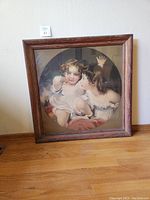 Full view of the framed print leaning against a wall on a wooden floor showing two children in a square wooden frame.