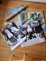 Four large art posters displayed on wooden floor with one shipping tube visible, showing the variety of subjects and color schemes in the lot.