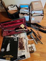 Overhead photo showing violin case partially open with bow and musical accessories inside, surrounded by boxes of sheet music and several music stands on wooden floor.