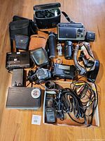 Full lot overview showing cameras, lenses, camera bags, flash unit, portable tape deck, and assorted cables and accessories spread on floor.