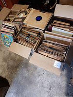 Six cardboard boxes filled with vinyl records and sleeves arranged on concrete floor