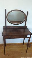 Full view of vintage wooden vanity with oval mirror mounted on two vertical supports and a drawer below the tabletop.