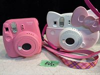 Photo showing two Fujifilm kids instant cameras side by side: a pink Instax Mini 9 and a white-pink Instax Mini Hello Kitty edition with themed bow and strap.