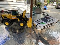 Photo showing both the yellow Tonka toy tractor with front loader and the white Tonka police car side by side on a glass table.