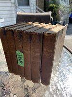 Photo of six antique brown hardcover books standing side by side on a glass table outdoors, titles on spines visible including Little Pitchers, Doctor Papa, Flaxie Frizzle, Flaxie Growing Up, Kittyleen, and The Twin Cousins.