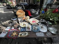 Photo showing a wide assortment of kitchen items including baskets, pots, pans, plastic and glass bowls, salt and pepper shakers, cake pan box, serving tray, trivets and more, laid out on a black surface with a car in the background.