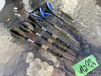 Photo of 5 complete brass metal darts with black and blue flights and 1 partially intact dart on glass table.