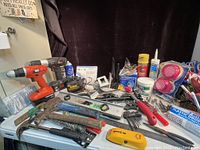 Full overview of assorted tools and hardware on white plastic table including cordless drills, hand tools, hardware, aerosol cans, gloves, masks, and small accessories.