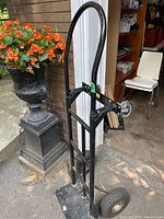 Front and side view of black metal appliance mover dolly with two large pneumatic wheels and two swivel casters, positioned outdoors near a wall and plant.