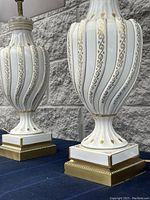Close-up of two white ceramic lamps with gold painted decorative swirls and metal bases with gold trim on a dark cloth surface in front of gray stone wall