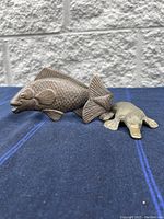 Two small brass fish figurines and a brass platypus figurine on blue surface against grey brick wall