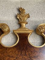 Close-up photo of the top decorative carved flame finial and floral scroll in gold finish on the wooden frame.