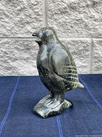 Full view of bird sculpture showing the body, carved wings, and standing base on blue cloth against stone wall background.