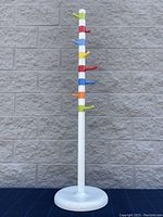 Front view of the colorful standing coat rack against a gray block wall, showing its design and condition.