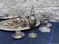 Photo showing silver-plated salt shaker, candlestick holders, and other small silver items on a blue cloth against a stone wall
