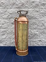 Photo showing full fire extinguisher body front view against white brick wall, copper body with brass label plate and red hose attached.