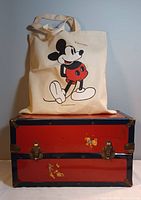 Photo of the off-white cotton tote bag with large Mickey Mouse graphic placed on top of the red and black vintage metal toy chest decorated with Winnie the Pooh decals.