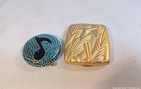 Photo of two Estee Lauder compacts, one round with light blue Swarovski crystals and black musical note, and one square with matte gold bamboo design and rhinestones
