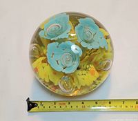 Top view of the large clear glass paperweight showing the blue flowers inside with yellow accents along with a tape measure for scale indicating about 5.5 inches diameter.