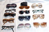 Twelve assorted sunglasses and prescription glasses arranged on white background, showing variety in frame shape and color.