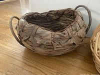 Large brown leaf basket with two loop handles on wooden floor.