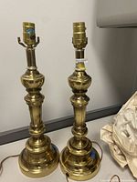 Two heavy brass table lamps with ornate columnar bodies and bulb sockets, shown side by side on a flat surface.
