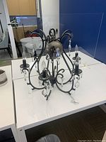 Photo of a black metal chandelier with five arms, each with candle-style light bulbs and hanging clear crystal pendants, shown on a white table in a room.
