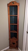 Solid wood corner cabinet standing in corner; natural wood frame with black interior and 3 shelves plus bottom cupboard door with heart cutout