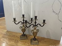 Photo showing pair of vintage metal candelabras with three candle holders each, in bronze-gold finish with detailed base, set on wooden floor against stone wall.