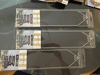 Three packs of long black event signs with white text and decorative corner borders, in clear plastic packaging with hang tabs.