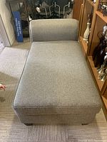 Photo of chaise lounge upholstered in light gray fabric with a curved backrest on one side, placed on carpet next to shelving unit, showing shape and size.
