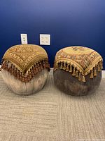 Two round poufs with fabric tops and tassels placed side by side on carpet with blue wall background