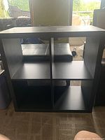 Front view of a single black modular storage cube showing four open compartments, placed indoors with a carpeted floor and partial background of furniture and outdoor view.