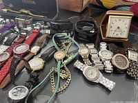 Over a dozen watches laid out on a table, showing various styles including analog watches with leather and silicone bands, metal bands, and a small foldable clock.