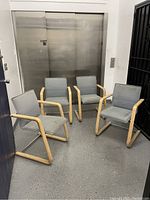 Set of four birch wood chairs with gray upholstered cushions placed in front of an elevator door on a gray floor.