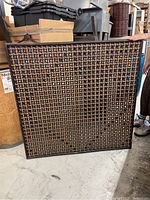 Square wooden decorative divider panel leaning against items in a storage area, shows front and framing with interwoven wooden grid pattern.