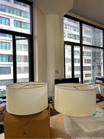 Two white fabric drum lampshades placed on cardboard boxes in a room with large windows, showing the exterior city view.