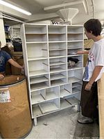Front view of large white metal mail room cabinet held by two people showing multiple compartments for sorting.