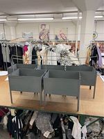 Two gray metal bins with legs and dividers, top front view. Background shows clothing racks.