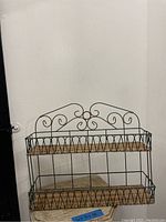 Front view of wicker and black metal spice rack showing two tiers with wicker bottom shelves, topped with decorative scroll metalwork