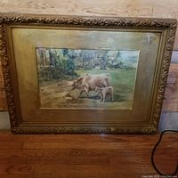 Photo of the antique framed painting of a cow and her calf placed against a wall, showing the detailed scenery and ornate frame.