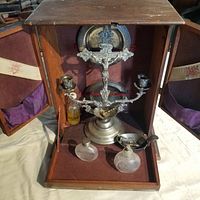 Open oak cabinet revealing purple velvet-lined interior housing silver-tone crucifix candelabra, two communion plates, glass bottles, brush and pourer