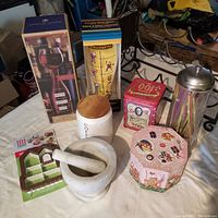 Photo showing all items in the lot on a white surface: boxed novelty wine glass, novelty beer glass box, two antique tins, coffee jar with wooden lid, gingerbread cookie cutter, glass straw holder with metal lid, and marble mortar and pestle.