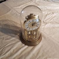 Battery operated anniversary clock inside clear glass dome with gold-tone base on fabric surface.