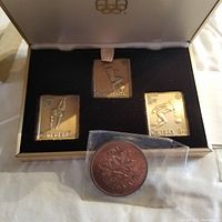 Three rectangular Canadian Olympic coins in a display box alongside a large 1965 penny in plastic.