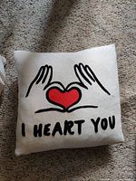 Cream decorative throw pillow with 'I HEART YOU' text and two hands holding a red heart graphic design on carpet background